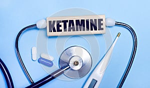 On a light gray background, a stethoscope, an electronic thermometer, pills, a wooden block with the text KETAMINE. Medical