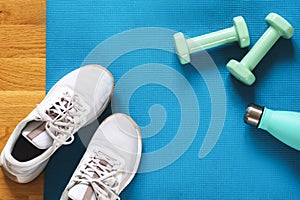 Light green dumbbells, bottle with water and white sneakers on blue sports mat background. Training at home concept. Top
