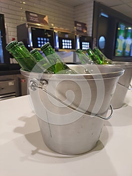 Light green glass beer bottles in a metal bucket with ice on a bar counter .