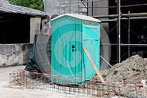 Light green portable ecological toilet left on construction site surrounded with sand and other construction material with old photo