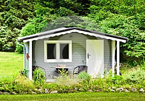 A light grey and white wooden small shed, gardenhouse, with a table and two chairs at the veranda