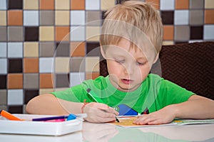 A light-haired boy in a green T-shirt is painting a coloring book while sitting at his desk at home. Correct organization of