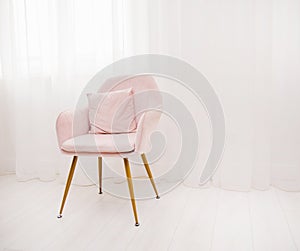A light pink armchair stands by the window against a background of white airy curtains. A lonely chair in the room