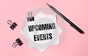 On a light pink background, black paper clips, black pen and white note paper with the words UPCOMING EVENTS. Flat lay