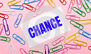 On a light pink background, colorful bright paper clips and a white card with the text CHANCE