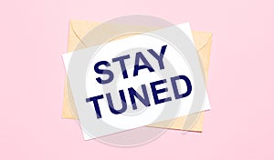 On a light pink background - a craft envelope. It has a white sheet of paper that says STAY TUNED