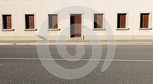 Light pink facade with row of closed wooden window blinds and a door in the middle. Sidewalk and urban street in front