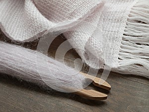 Light pink fringed shawl made of fluffy mohair yarn. Ready handwoven garment and shuttle with thread on wooden table