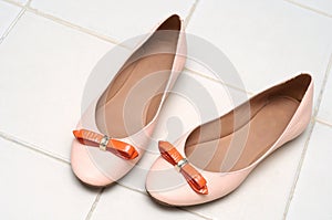 A light pink pinkish ladies shoes with bold orange colored ribbon on a beige ceramic tiled floor