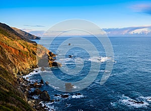 The light of the setting sun illuminates the reds, greens, and browns of the steep mountains and cliffs of the Big Sur coast