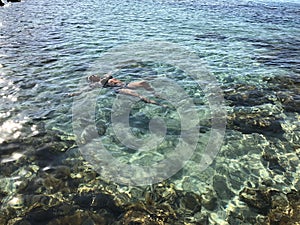 Light-skinned girl in black swimsuit swims in the clear sea with colored stones and turquoise water, top view