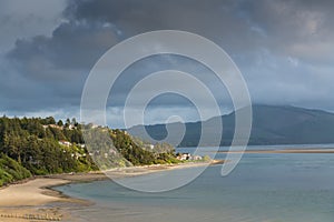 Light from sunset spotlights homes and a golden sandy beach along a calm bay under dramatic clouds photo