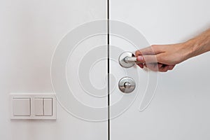 Light switch next to the door handle. Open white door with metallic handle. The man opens the door.