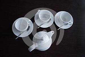 Light tea dishes are placed on a dark table. A white teapot and three mugs with saucers and spoons