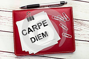 On a light wooden background, a burgundy pen and notebook. On the notebook has white paper clips and white paper with the text