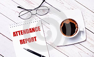 On a light wooden background, glasses, a pen, a cup of coffee and a notepad with the text ATTENDANCE REPORT