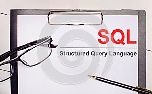 On a light wooden background glasses, a pen and a sheet of paper with the text SQL Structured Query Language. Business concept