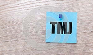 On a light wooden background - a light blue square sticker with the text TMJ