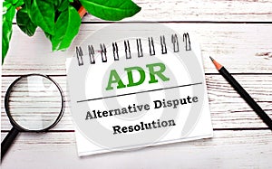 On a light wooden background, a magnifying glass, a pencil, a green plant and a white notebook with text ADR Alternative Dispute