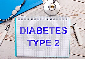 On a light wooden background there is a stethoscope, a blue notebook, a white pen and a sheet of paper with the text DIABETES TYPE