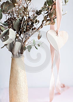 Light wooden heart pendant on a pink silk ribbon on a background of a golden shiny glittered vase with dried eucalyptus branches