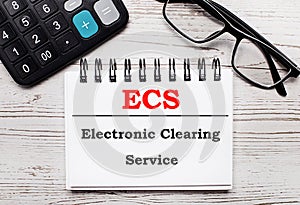 On a light wooden table calculator, glasses and a blank notepad with the text ECS Electronic Clearing Service. Business concept