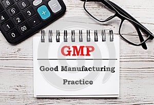 On a light wooden table calculator, glasses and a blank notepad with the text GMP Good Manufacturing Practice. Business concept