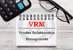 On a light wooden table calculator, glasses and a blank notepad with the text VRM Vendor Relationship Management. Business concept