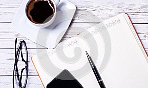 On a light wooden table is an open notebook with a pen, a smartphone, black-rimmed glasses and a cup of coffee. Workplace close up