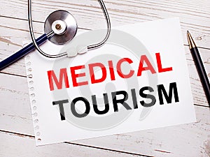 On a light wooden table there is a stethoscope, a pen and a sheet of paper with the text MEDICAL TOURISM. Medical concept
