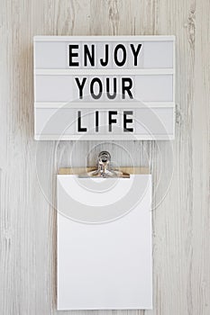 Lightbox with text `Enjoy your life`, noticepad with blank sheet of paper on a white wooden background, top view. Overhead, from