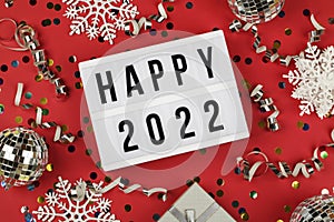 lightbox with words happy 2022, gift box and holiday confetti on red background. top view new year celebration.