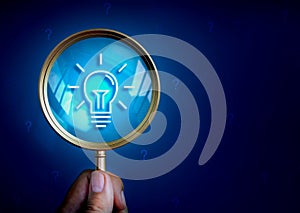 Lightbulb icon in magnifying glass lens in hand on dark blue background with many question mark symbol.