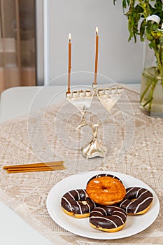 Lighted Hanukkah with one candle and shamash on the first day of Hanukkah on the festive table next to donuts and a vase