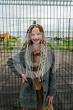 Lighthearted teenage girl with dreads standing in front of a metal fence