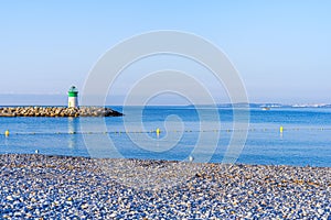 Lighthouse on the background of the sea and the beach of small stones, the sea is full calm and clear. Blue sky, summer