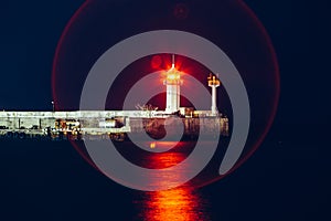 Lighthouse with red light reflected in night black sea water on end of pier in Yalta, Crimea