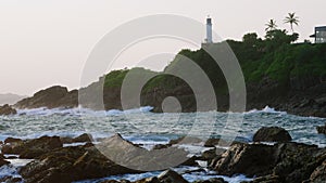 Lighthouse stands on green hill above ocean waves crashing against rocky shore at sunset. Tranquil dynamic nature