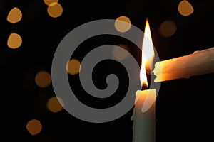 Lighting candle from another one against blurred lights in darkness, closeup. Space for text