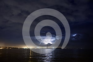 Lightning above the sea or lake at night. Thunderstorm above large industrial city skyline.