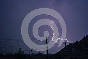 A lightning bolt strikes down to earth in a romanian village with power pole and power lines in the foreground.