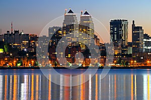 The lights of the Beautiful tall buildings, skyscrapers, towers are reflected in the Dnieper River at sunset in the evening.