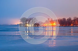 Lights of evening city lanterns. View of the embankment and the bridge to the island from the surface of the frozen lake. Island