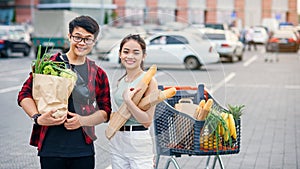 Likable asian couple hold paper eco bags with organic healthy food in hands while standing near store mall. Happy family