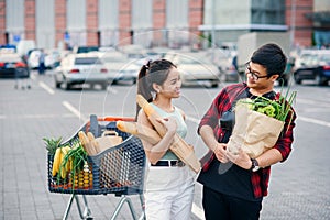 Likable asian woman and handsome vietnamese young man look to each other holding paper eco bags with organic healthy