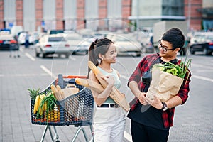 Likable asian woman and handsome vietnamese young man look to each other holding paper eco bags with organic healthy photo