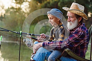 Likable respected 70-aged bearded grandfather holding on knees 10-aged cute grandson and teaching him to catch fish.