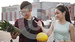 Likable smiling asian man weighting in hands peanapple and melon and his pretty girlfriend trying to guess what is