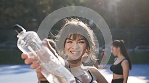 Likable smiling slender girl posing on camera with bottle of water after fitness lesson outdoors