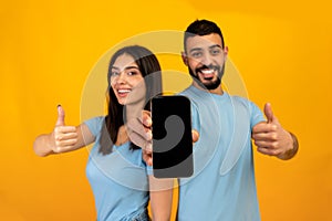 We like this app. Young arab couple showing smartphone blank screen and gesturing thumbs up, yellow background, mockup